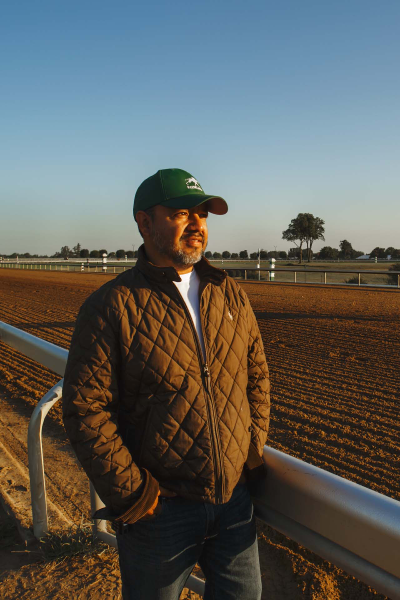A photo of Alfredo Laureano looking off into the distance while standing by the dirt track at sunset. He is an older Hispanic man with graying chin stubble. He is wearing a green Keencanter-brand hat and a brown Keencanter-brand jacket overtop a white shirt.