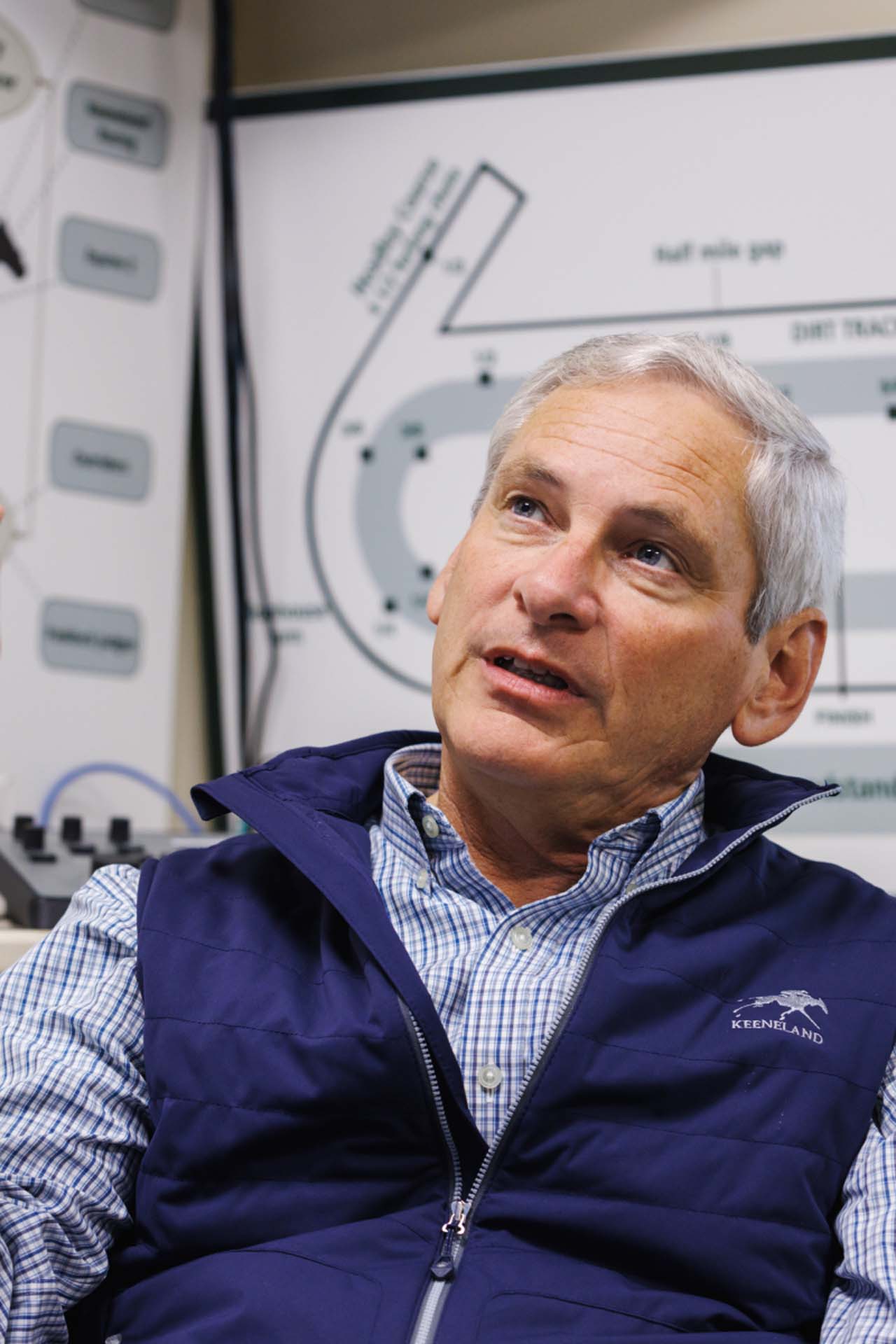 An up-close photo of Dr. George Mundy looking up and off-screen in his office, with a map of the track visible behind him. He is an older White man with short silver hair. He is wearing a navy Keencanter-brand vest over a white and blue checkered shirt.